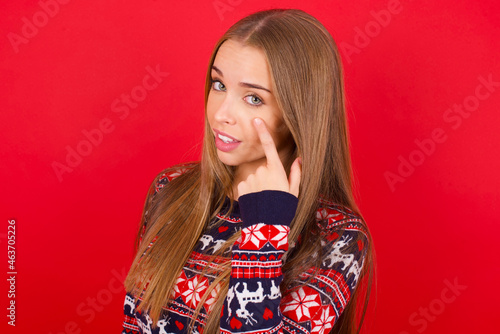 Young caucasian girl wearing christmas sweaters on red background Pointing to the eye watching you gesture, suspicious expression.