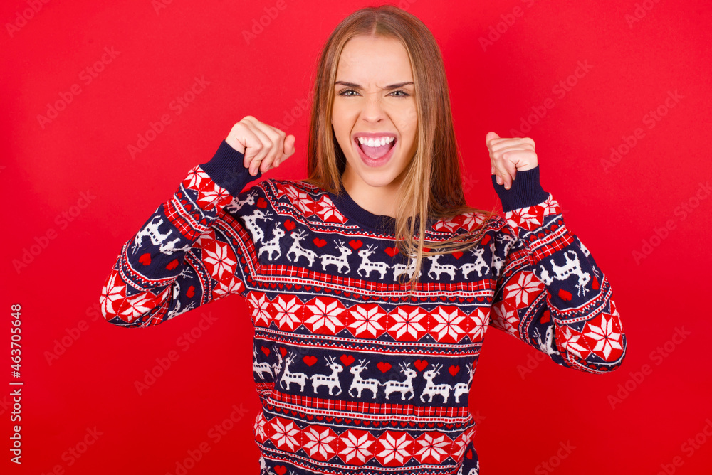 Young caucasian girl wearing christmas sweaters on red background celebrating surprised and amazed for success with arms raised and open eyes. Winner concept. Young caucasian girl wearing christmas sweaters on red background celebrating surprised and amazed for success with arms raised and open eyes. Winner concept.