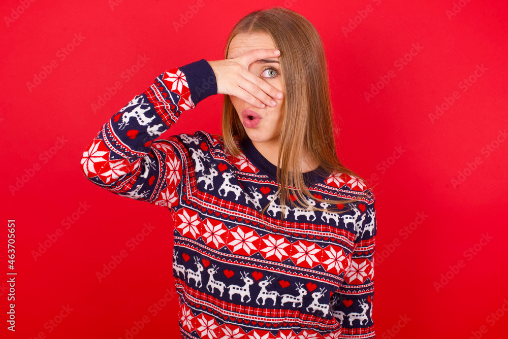 Young caucasian girl wearing christmas sweaters on red background peeking in shock covering face and eyes with hand, looking through fingers with embarrassed expression. Young caucasian girl wearing christmas sweaters on red background peeking in shock covering face and eyes with hand, looking through fingers with embarrassed expression.