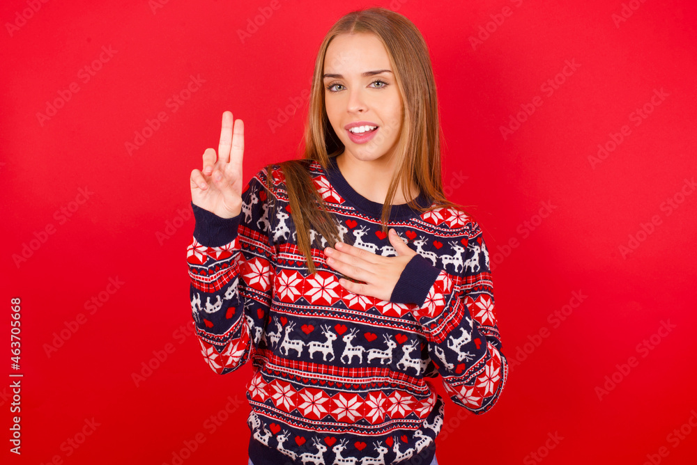 Young caucasian girl wearing christmas sweaters on red background smiling swearing with hand on chest and fingers up, making a loyalty promise oath. Young caucasian girl wearing christmas sweaters on red background smiling swearing with hand on chest and fingers up, making a loyalty promise oath.