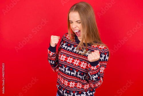 Young caucasian girl wearing christmas sweaters on red background very happy and excited doing winner gesture with arms raised, smiling and screaming for success. Celebration concept.