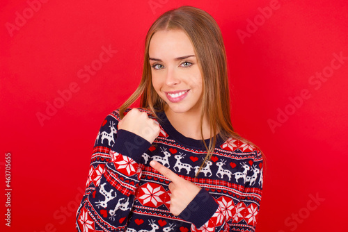 Young caucasian girl wearing christmas sweaters on red background In hurry pointing to wrist watch, impatience, looking at the camera with relaxed expression