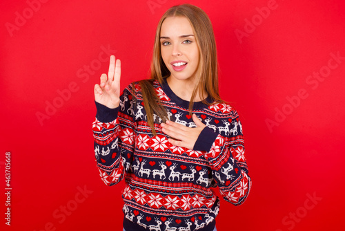 Young caucasian girl wearing christmas sweaters on red background smiling swearing with hand on chest and fingers up, making a loyalty promise oath.