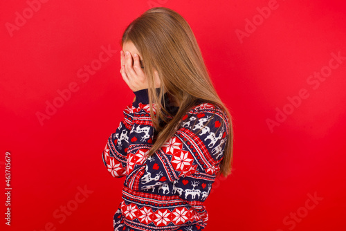 Sad Young caucasian girl wearing christmas sweaters on red background crying covering her face with her hands.