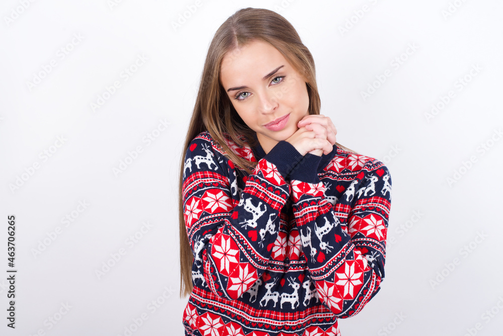 Charming serious Young caucasian girl wearing christmas sweaters on white background keeps hands near face smiles tenderly at camera