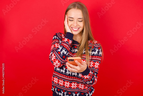 Young caucasian girl wearing christmas sweaters on red background holding in hands cell reading sms using new app 5g