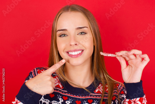 Fototapeta Naklejka Na Ścianę i Meble -  Young caucasian girl wearing christmas sweaters on red background holding an invisible aligner and pointing to her perfect straight teeth. Dental healthcare and confidence concept.