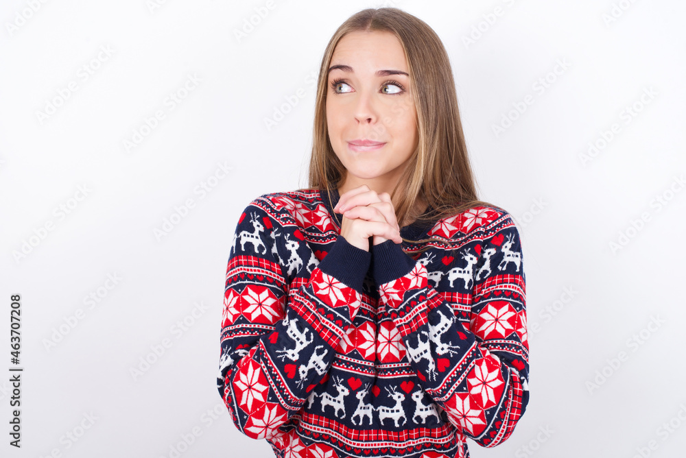 Curious Young caucasian girl wearing christmas sweaters on white background keeps hands under chin bites lips and looks with interest aside. Curious Young caucasian girl wearing christmas sweaters on white background keeps hands under chin bites lips and looks with interest aside.