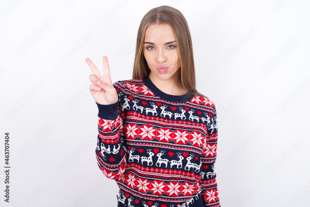 Young caucasian girl wearing christmas sweaters on white background makes peace gesture keeps lips folded shows v sign. Body language concept Young caucasian girl wearing christmas sweaters on white background makes peace gesture keeps lips folded shows v sign. Body language concept