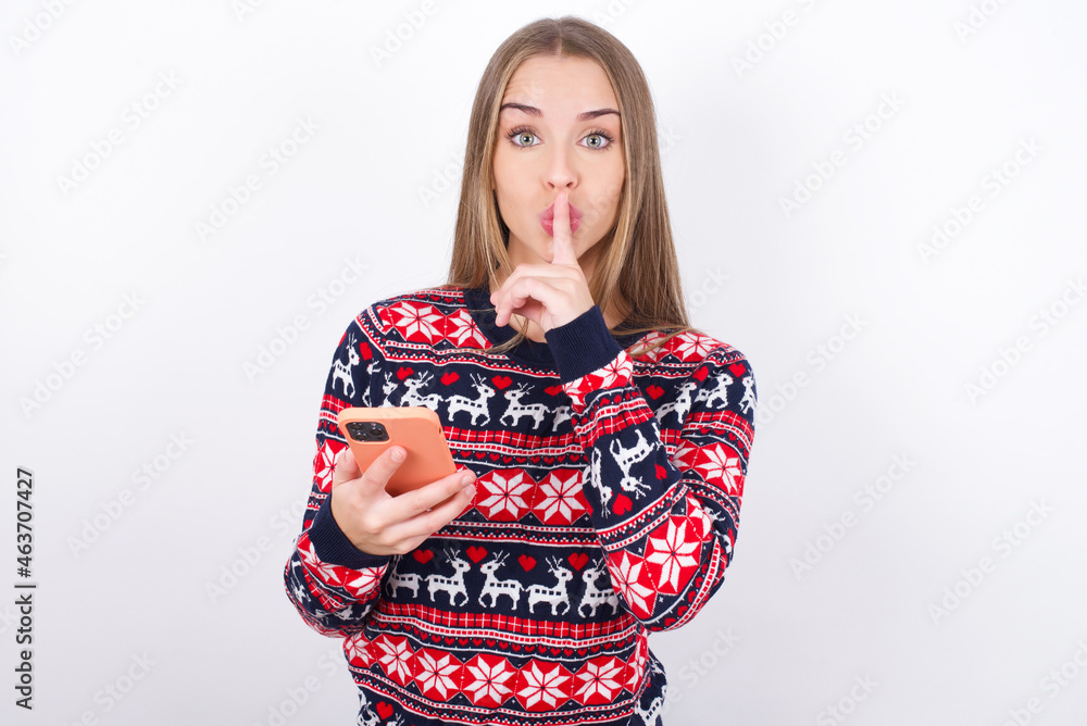 Young caucasian girl wearing christmas sweaters on white background holding modern gadget ask not tell secrets Young caucasian girl wearing christmas sweaters on white background holding modern gadget ask not tell secrets