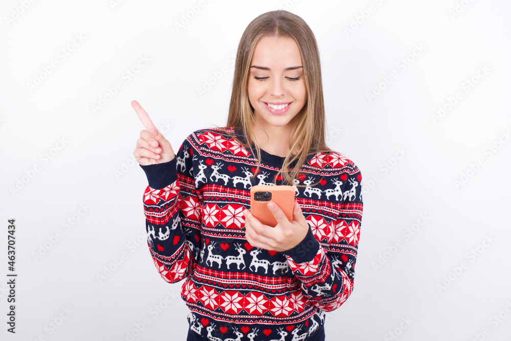 Smiling Young caucasian girl wearing christmas sweaters on white background pointing finger at blank space holding phone in one hand Smiling Young caucasian girl wearing christmas sweaters on white background pointing finger at blank space holding phone in one hand