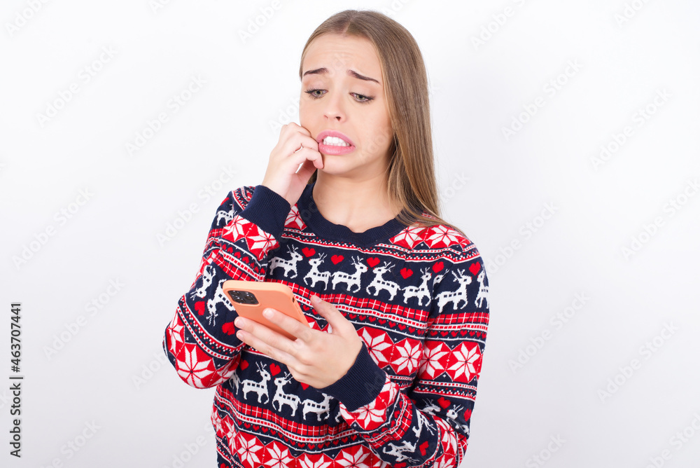Portrait of pretty frightened Young caucasian girl wearing christmas sweaters on white background chatting biting nails after reading some scary news on her smartphone. Portrait of pretty frightened Young caucasian girl wearing christmas sweaters on white background chatting biting nails after reading some scary news on her smartphone.