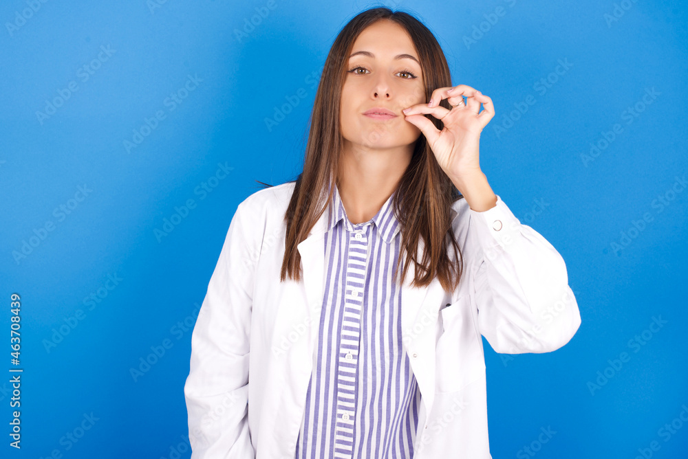Young european doctor woman on blue background mouth and lips shut as zip with fingers. Secret and silent, taboo talking. Young european doctor woman on blue background mouth and lips shut as zip with fingers. Secret and silent, taboo talking.