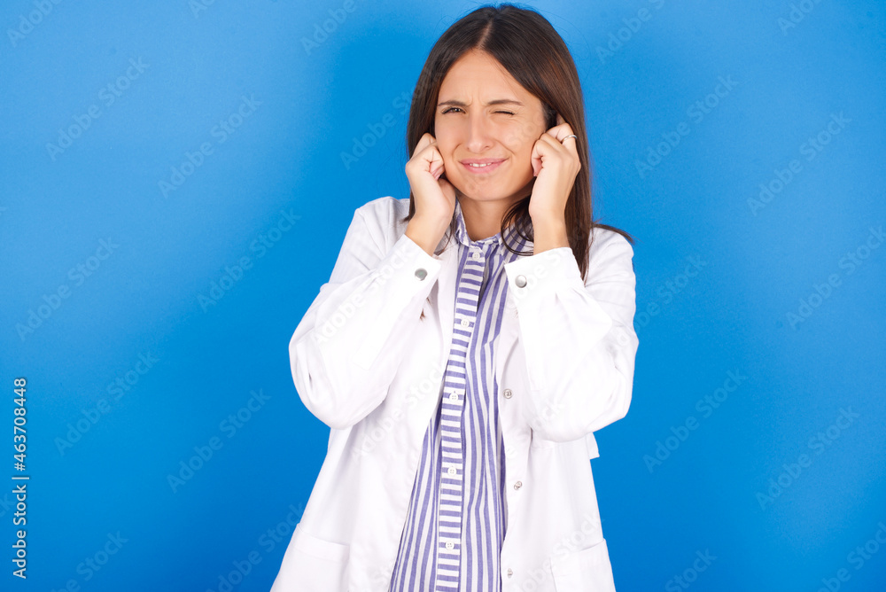 Young european doctor woman on blue background covering ears with fingers with annoyed expression for the noise of loud music. Deaf concept. Young european doctor woman on blue background covering ears with fingers with annoyed expression for the noise of loud music. Deaf concept.