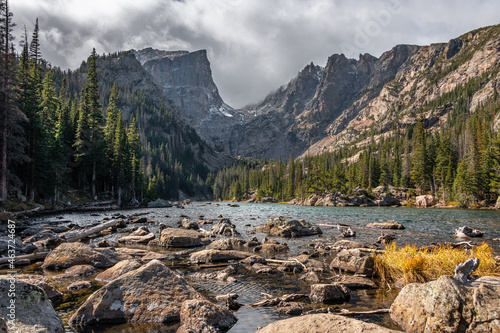 View of dream lake surrounded by mountains in rocky mountain national park, colorado. The lake has many rocks and the peak of the mountain is pointed and surrounded by trees.