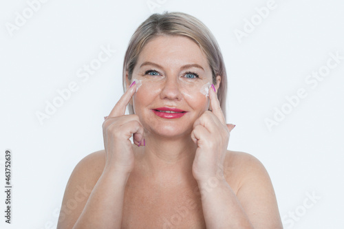 Happy 40s mid aged mature blonde lady applying facial cream on face looking at camera isolated on white background. Anti age healthy dry skin care beauty