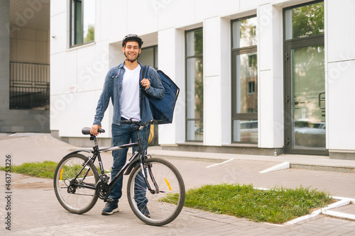Portrait of joyful handsome young delivery man in protective helmet posing standing near bicycle in city street, looking at camera. Courier male with thermo backpack delivery food to client
