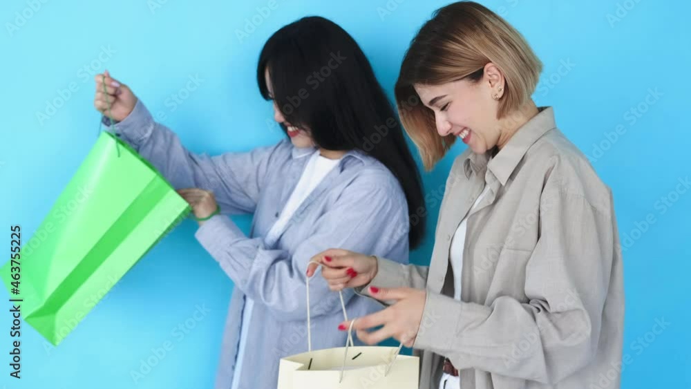 Friends shopping. Delivery service. Female leisure. Online store. Excited happy interracial Asian Caucasian girls best buddies receiving bags order isolated on blue background. Friends shopping. Delivery service. Female leisure. Online store. Excited happy interracial Asian Caucasian girls best buddies receiving bags order isolated on blue background.