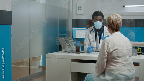 Young medic wearing face mask while consulting elder patient behind plexiglass wall at medical facility. Doctor helping senior woman with healing treatment for healthcare during pandemic