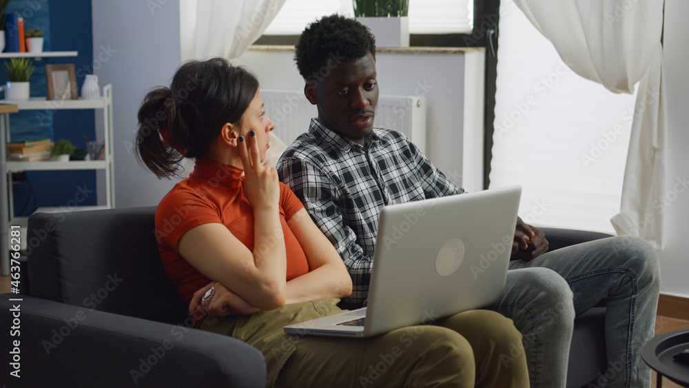 Married couple of interracial partners looking at laptop and talking while sitting on sofa in living room. Multi ethnic family using modern device discussing lifestyle at home with technology Married couple of interracial partners looking at laptop and talking while sitting on sofa in living room. Multi ethnic family using modern device discussing lifestyle at home with technology