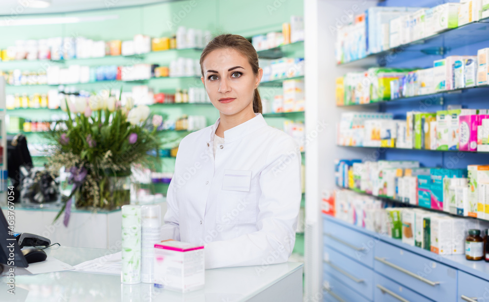 Portrait of cheerful positive smiling woman pharmacist who is standing on her work place near cashbox in apothecary.