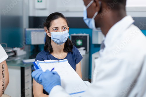 Closeup of african american pediatrician doctor writing ill expertise on clipboard explaining healthcare treatment to parent during clinical examination in hospital office. Therapist with face mask