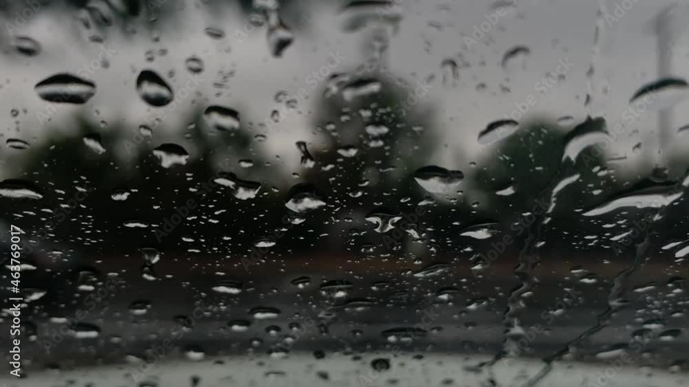 Close up of a window with rain drops falling down, Heavy rain on window. Rain splashing on a window, over a car passing and blurred tree and sky in the park outside the car. Close up of a window with rain drops falling down, Heavy rain on window. Rain splashing on a window, over a car passing and blurred tree and sky in the park outside the car.