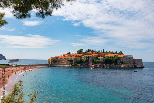 A picturesque panoramic view of the island of Sveti Stefan, the orange roofs of the island hotel in the green of cypress trees, the azure transparent sea. On the beach, people sunbathe and swiming
