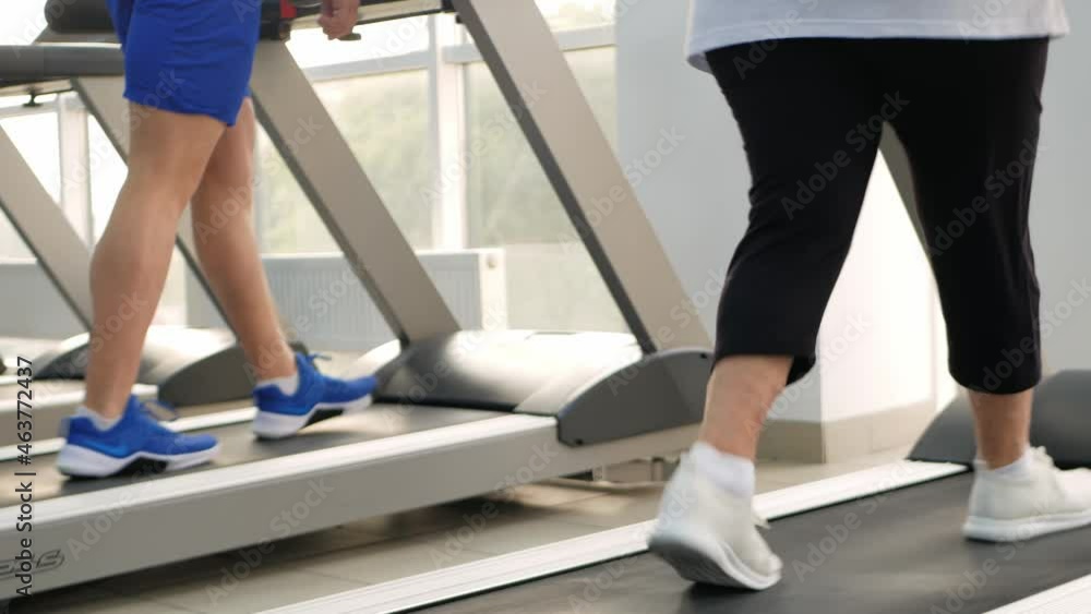 Happy smiling people are engaged in physical education, simulators in the gym. Active healthy lifestyle, senior concept. A woman with gray hair, a young coach is a muscular man. feet on the treadmill Happy smiling people are engaged in physical education, simulators in the gym. Active healthy lifestyle, senior concept. A woman with gray hair, a young coach is a muscular man. feet on the treadmill