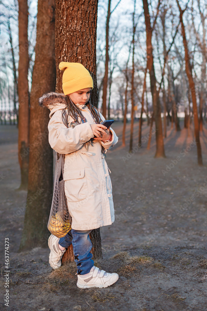 Teenager girl uses a smartphone on her way home through the autumn park after shopping, bright yellow fruits in a gray string bag, eco mesh with purchase, zero waste and sustainable lifestyle