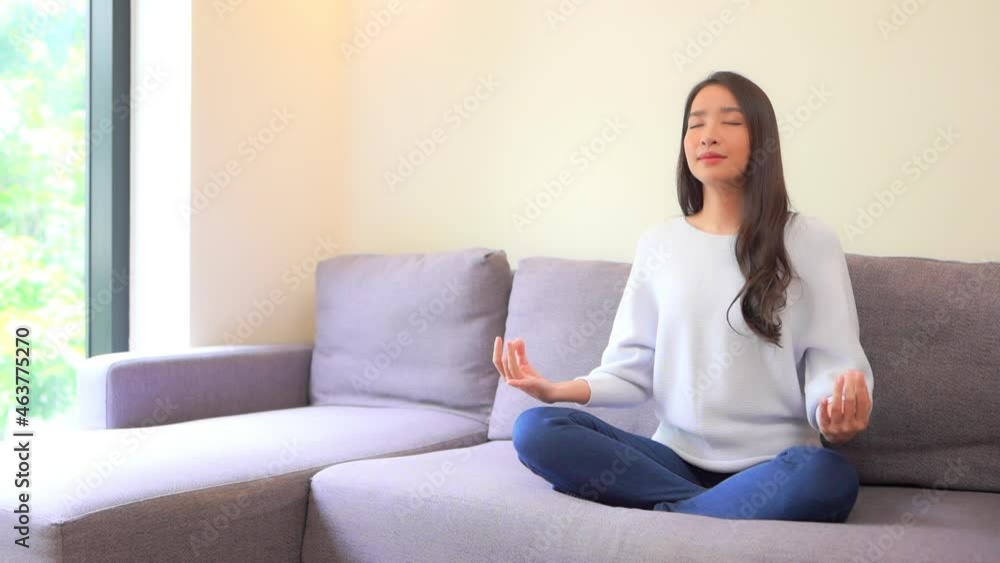 Asian young woman meditating on her sofa in the living room near a window. Asian young woman meditating on her sofa in the living room near a window.