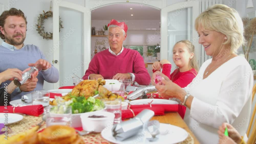 Multi generation family pulling Christmas crackers before putting on paper hats and eating meal at home together - shot in slow motion Multi generation family pulling Christmas crackers before putting on paper hats and eating meal at home together - shot in slow motion