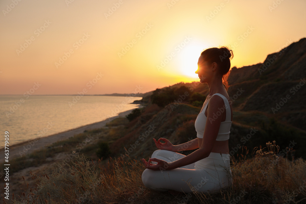 Mature woman meditating on hill near sea at sunset. Space for text