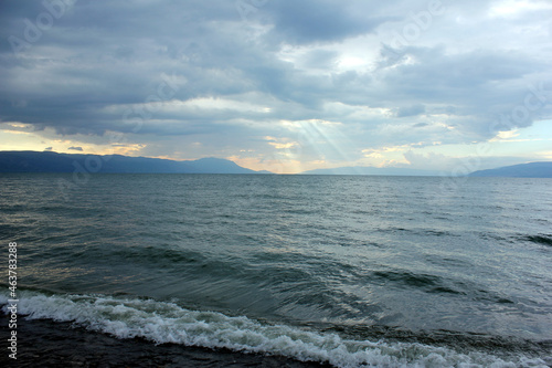 Dramatic cloudy sky with shades and lines of sunlight dropping on waves on the sea and mountains on the far background.