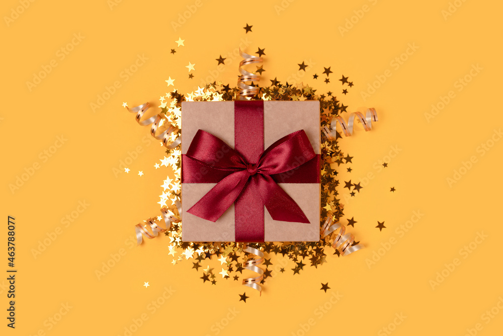 Gift box with red ribbon and gold confetti on a yellow background. Gift box with red ribbon and gold confetti on a yellow background.