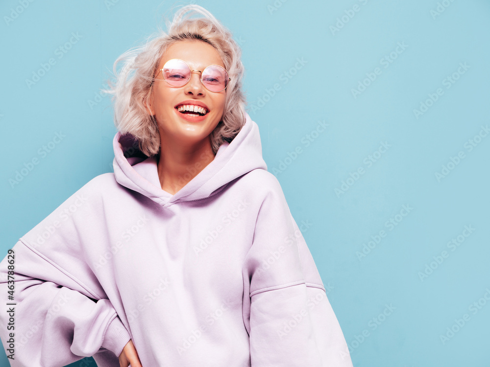 Young beautiful smiling female in trendy summer hoodie. Sexy carefree woman posing near blue wall in studio. Positive blond model having fun and going crazy. Cheerful and happy. In sunglasses Young beautiful smiling female in trendy summer hoodie. Sexy carefree woman posing near blue wall in studio. Positive blond model having fun and going crazy. Cheerful and happy. In sunglasses