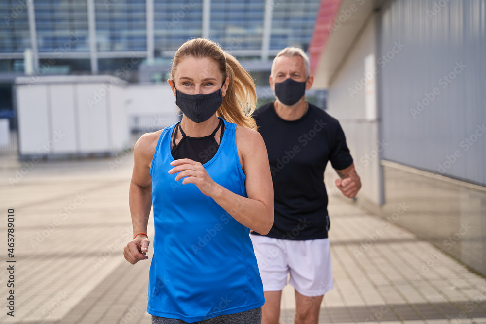 Focused middle aged woman wearing protective mask while running together with her husband outdoors in the morning to stay fit during Covid19 pandemic