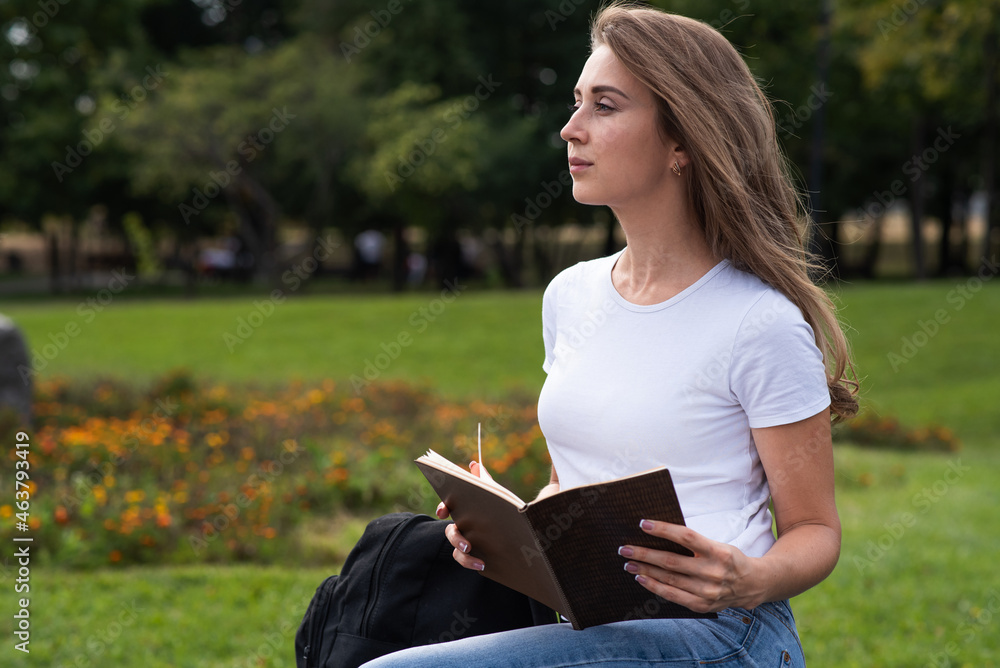 Obraz premium A young, happy and attractive Caucasian girl is sitting in the park and reading a book. Education concept.