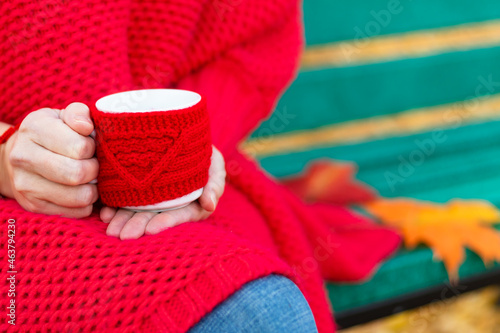 Female hands in a bright red sweater and bracelet hold a red mug with a hot drink on the background of a bench with autumn yellow leaves. Selective focus. Close-up