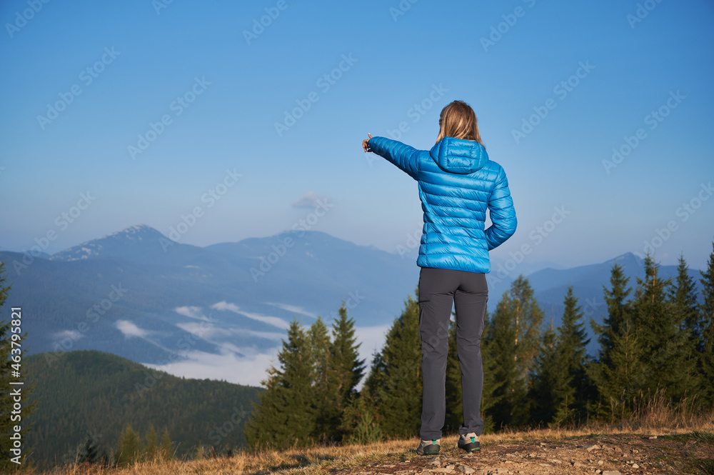 Back view of young woman hiker standing on hill and pointing finger at majestic mountains under blue sky. Female traveler gesturing towards foggy hills while enjoying beauty of nature in mountains. Back view of young woman hiker standing on hill and pointing finger at majestic mountains under blue sky. Female traveler gesturing towards foggy hills while enjoying beauty of nature in mountains.
