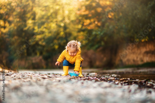 Autumn season. A cute little girl in yellow rubber boots is playing with a paper boat by the river. Sunny park on the background