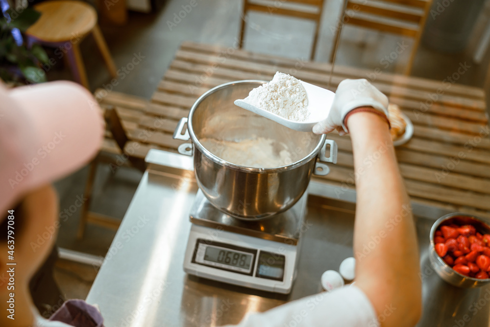 Baker pours white flour into container to weigh on digital scales in workshop Baker pours white flour into container to weigh on digital scales in workshop
