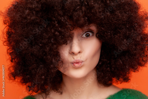 Closeup portrait of woman with Afro hairstyle looking at camera with pout lips, having perfect skin and fluffy hair, advertising beauty salon. Indoor studio shot isolated on orange background.