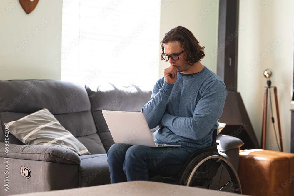 Thoughtful caucasian disabled man wearing glasses sitting on wheelchair using laptop at home Thoughtful caucasian disabled man wearing glasses sitting on wheelchair using laptop at home