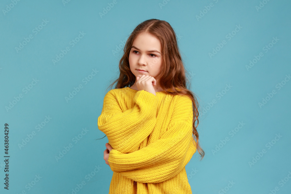 Portrait of pensive little girl stands looking away with hand on chin, thinks about important things, wearing yellow casual style sweater. Indoor studio shot isolated on blue background. Portrait of pensive little girl stands looking away with hand on chin, thinks about important things, wearing yellow casual style sweater. Indoor studio shot isolated on blue background.