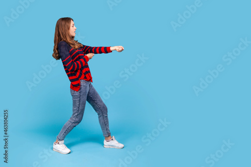 Full length profile portrait of woman wearing striped casual style sweater, standing in attack or pulling hands gesture and looking forward. Indoor studio shot isolated on blue background.