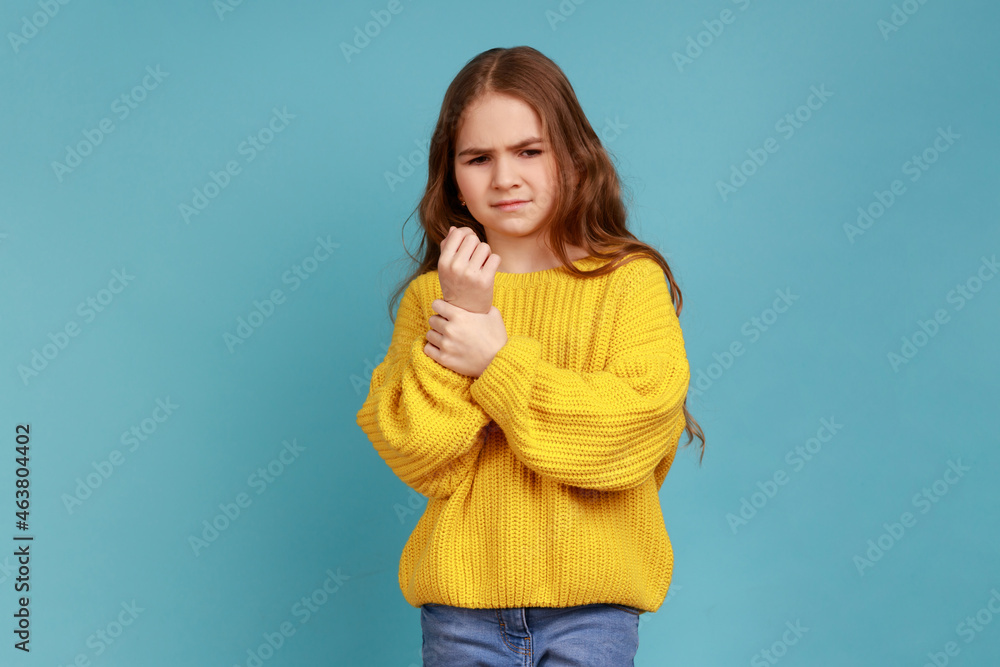 Portrait of little girl standing with grimace of pain, touching sore sprained wrist, health problem, wearing yellow casual style sweater. Indoor studio shot isolated on blue background. Portrait of little girl standing with grimace of pain, touching sore sprained wrist, health problem, wearing yellow casual style sweater. Indoor studio shot isolated on blue background.