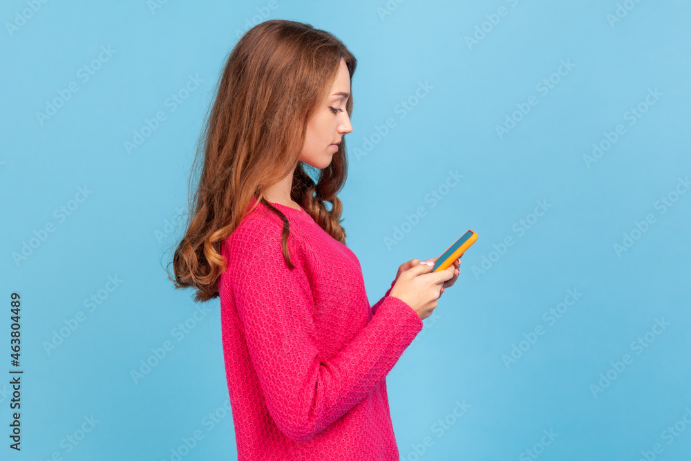 Side view of serious woman in pullover, typing on cell phone, reading blog, chatting in social network, using mobile app for online communication. Indoor studio shot isolated on blue background. Side view of serious woman in pullover, typing on cell phone, reading blog, chatting in social network, using mobile app for online communication. Indoor studio shot isolated on blue background.