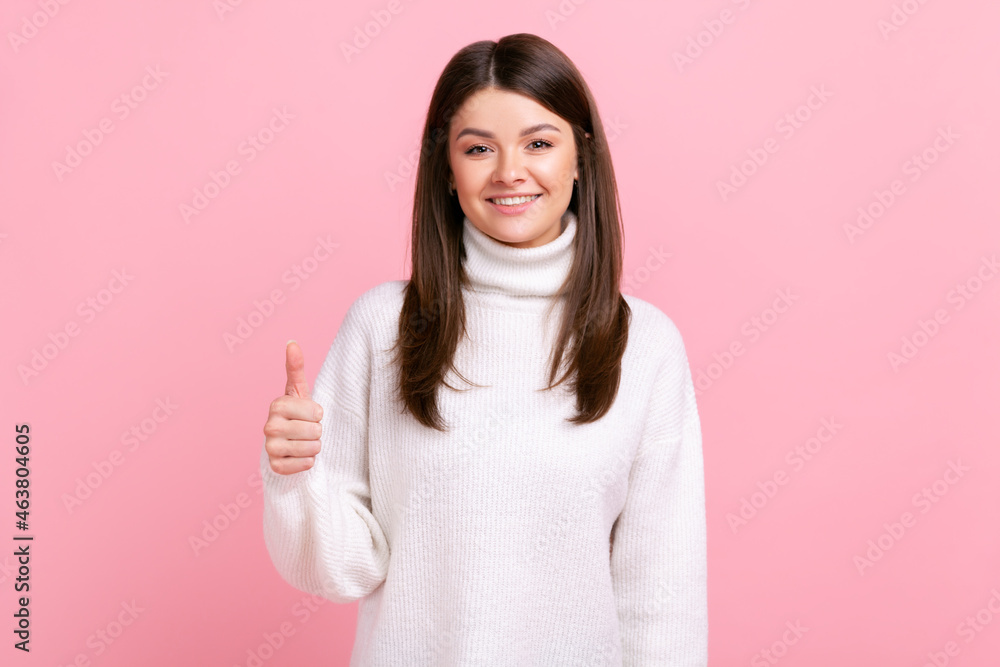 Smiling brunette female showing thumb up to camera, expressing approbation and positive emotions, wearing white casual style sweater. Indoor studio shot isolated on pink background. Smiling brunette female showing thumb up to camera, expressing approbation and positive emotions, wearing white casual style sweater. Indoor studio shot isolated on pink background.