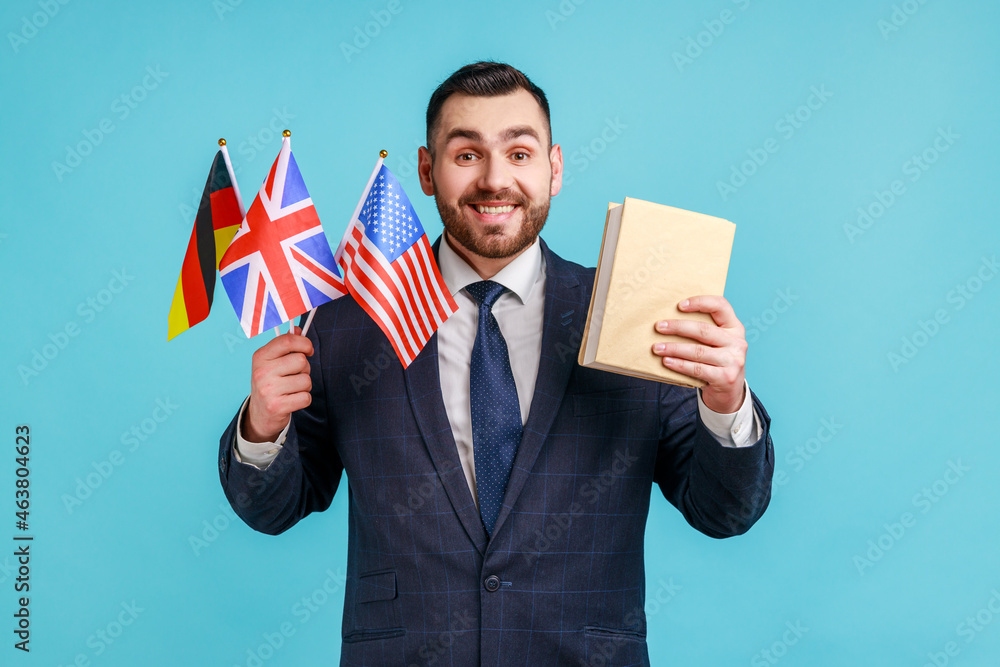 Satisfied businessman or teacher wearing official style suit, holding flags of Germany, Great Britain and USA and book, looking at camera with smile. Indoor studio shot isolated on blue background. Satisfied businessman or teacher wearing official style suit, holding flags of Germany, Great Britain and USA and book, looking at camera with smile. Indoor studio shot isolated on blue background.